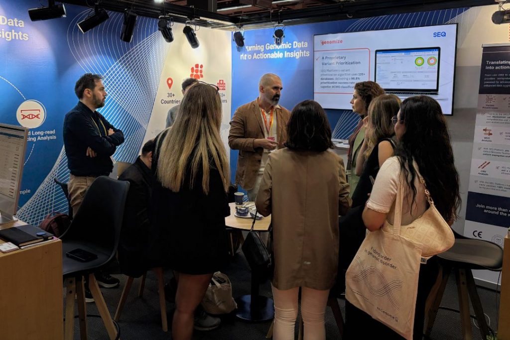 Visitors gather at the Genomize booth during a genomics conference, discussing the SEQ Platform and its genomic data analysis capabilities while viewing a live product demonstration on a presentation screen.