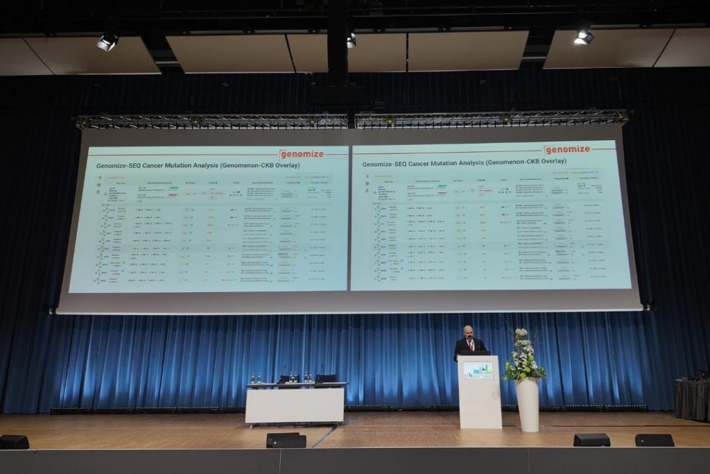 A speaker presents on stage at a scientific conference in a large auditorium. Two projection screens display Genomize SEQ Platform slides focused on cancer mutation analysis and Genomenon–CKB integration. The stage features a podium, conference seating, and blue curtain backdrops, with attendees and panel members visible in the presentation area.