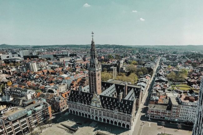 Genomize – ISV Aerial view of Leuven, Belgium, featuring the historic Gothic-style Town Hall and the iconic University Library tower at the city center. The image captures the surrounding urban landscape with red-roofed buildings, streets, and green spaces extending toward the horizon under a clear sky. The photograph provides a panoramic perspective of Leuven’s historic architecture and cityscape, the host city of the International Symposium on Variants in the Genome (ISV 2026).