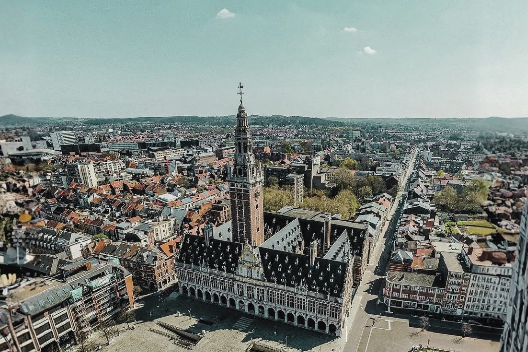 Aerial view of Leuven, Belgium, featuring the historic Gothic-style Town Hall and the iconic University Library tower at the city center. The image captures the surrounding urban landscape with red-roofed buildings, streets, and green spaces extending toward the horizon under a clear sky. The photograph provides a panoramic perspective of Leuven’s historic architecture and cityscape, the host city of the International Symposium on Variants in the Genome (ISV 2026).