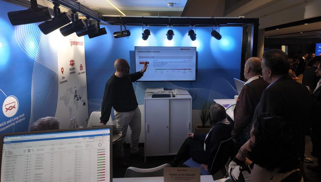 A Genomize team member demonstrates the SEQ Platform to visitors at a conference exhibition booth. Several attendees gather around a large display screen showing automated ACMG classification features, while branded booth graphics highlight global usage statistics, sample numbers, and genomic analysis capabilities. The image captures an interactive product demonstration and audience engagement during the event.