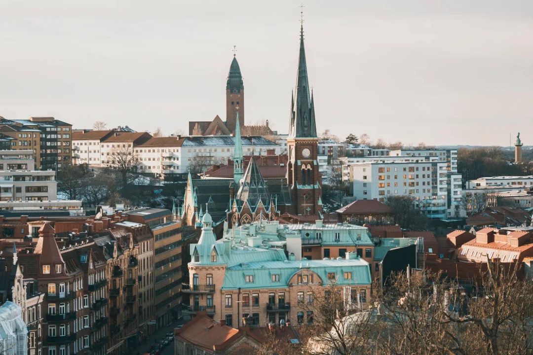 Aerial view of Gothenburg, Sweden, featuring historic church spires, red-roofed buildings, and a mix of classic and modern architecture. The cityscape highlights prominent landmarks rising above the urban skyline, with residential and commercial buildings extending into the distance. This image captures the character of Gothenburg, the host city of the European Human Genetics Conference (ESHG 2026).